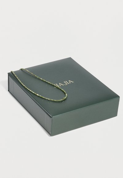 Σκουρόχρονο πράσινο κουτί κοσμημάτων με χρυσά γράμματα, στο οποίο βρίσκεται ένα λεπτό πράσινο κολιέ με χάντρες.