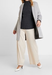 Šedý vlněný kabát, tmavý pletený svetr, krémové kalhoty s širokými nohavicemi a bílé lodičky s špičatou špičkou. Hladké textury, přizpůsobený střih a vyvážená barevná paleta.