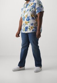 Lyhythihainen valkoinen paita, jossa sinisiä ja keltaisia kukkakuoseja, yhdistetty sinisiin farkkuihin ja valkoisiin tennareihin. Henkilö seisoo käsi rentona.