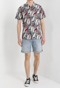Camisa de manga corta con estampados rojos y verdes sobre un fondo crema, combinada con pantalones cortos de mezclilla claros y zapatillas negras.