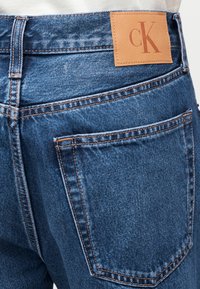 Κοντινή λήψη πίσω τσέπης μπλε τζιν με καφέ δερμάτινο ταμπελάκι με το λογότυπο "cK" και πορτοκαλί ραφές λεπτομέρειας.