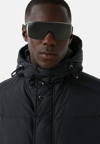 Schwarze Steppjacke mit hohem Kragen, Knopfverschlüssen und Kordelzügen. Dunkle Sonnenbrille mit breiten Gläsern und schlankem Design.