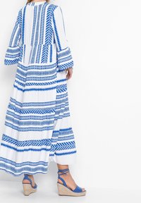 Robe maxi bleue et blanche à motifs avec manches longues, présentant des designs géométriques ; associée à des sandales à talons compensés bleues et à semelle texturée.