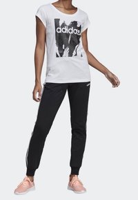 Vit t-shirt med svart Adidas-logotyp och grafisk tryck, kombinerad med svarta joggerbyxor med vita ränder och elastiska mudd, samt rosa sneakers.