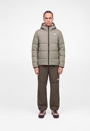Jeune homme aux cheveux tressés debout, portant une doudoune grise, un pantalon cargo olive et des chaussures de randonnée beige sur un fond uni.