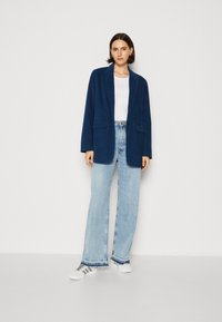 Meri blazer corduroy biru laut dengan satu kancing, dua saku depan, dipadukan dengan celana jeans lebar biru muda dan sepatu sneakers putih.