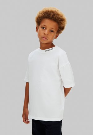 Ung dreng med krøllet hår iført en ensfarvet hvid Calvin Klein T-shirt og sorte bukser, stående foran en grå baggrund.