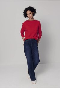 Punainen neulepaita, siniset leveälahkeiset farkut ja valkoiset lenkkitossut. Tausta on yksinkertainen harmaa. Malli seisoo rennosti käsivarret alhaalla ja ilme neutraali.