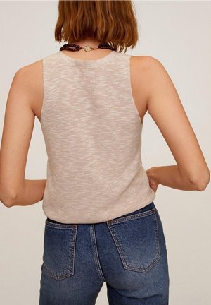 Femme aux cheveux courts portant un haut en maille beige sans manches et un jean en denim bleu, vue de dos sur un fond neutre.