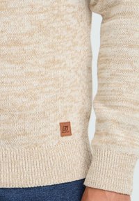 Beige strikket genser med ermer og nedre del som har ribbestrikkede mansjetter, samt et lite brunt lærmerke over blå denimjeans.