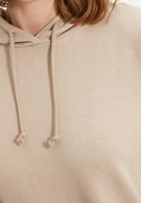 Sudadera con capucha beige, hecha de un tejido suave y texturizado, con un cordón ajustable con extremos anudados y un corte relajado.