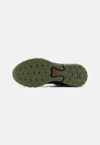 Semelle d’une chaussure de trail verte et noire avec des crampons profonds et un petit logo rouge au centre.