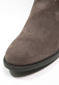 Bout de chaussure en daim marron avec semelle en caoutchouc noire et coutures visibles sur fond blanc.