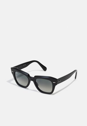Gafas de sol negras con un diseño cuadrado, lentes de gradiente gris y sutiles acentos plateados en la montura. Fabricadas en un material plástico ligero.