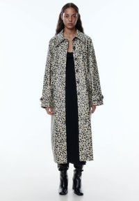Leopardenprint lange jas van glanzende stof met een kraaglijn, knoopsluiting en opgerolde mouwen, gecombineerd met een zwarte jurk en laarzen.