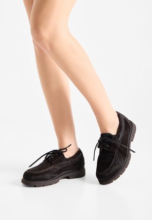 Gambe con scarpe stringate nere pelose con suole spesse, in piedi su uno sfondo bianco uniforme.