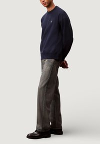 Felpa blu navy con scollatura rotonda e polsini a costine, abbinata a jeans grigio chiaro a gamba larga e scarpe in pelle nere.