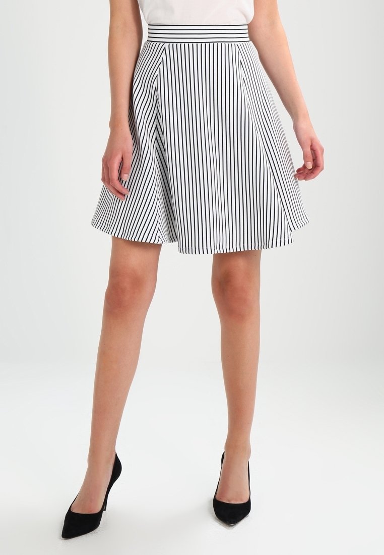 Femme portant une jupe blanche à rayures noires verticales, associée à un haut blanc et des chaussures à talons noirs, debout sur un fond uni.