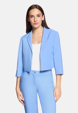 Kobieta ubrana w jasnoniebieską, krótką marynarkę i dopasowane spodnie, na białym tle, stojąca na jednolitym, jasnym tle.