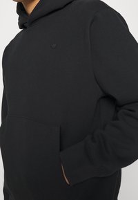 Svart huvtröja i mjukt tyg med en framficka. Har en liten, präglad logotyp på övre vänstra sidan. Muddar vid ärmar och huva.