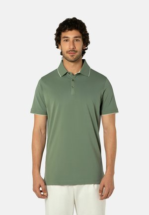 Man staat frontaal en draagt een lichtgroen poloshirt met korte mouwen en een witte broek tegen een egale lichte achtergrond.