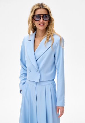 Kobieta o długich blond włosach, nosząca przyciemniane, duże okulary przeciwsłoneczne, złote kolczyki koła, jasnoniebieską krótka marynarkę i dopasowane spodnie z wysokim stanem.