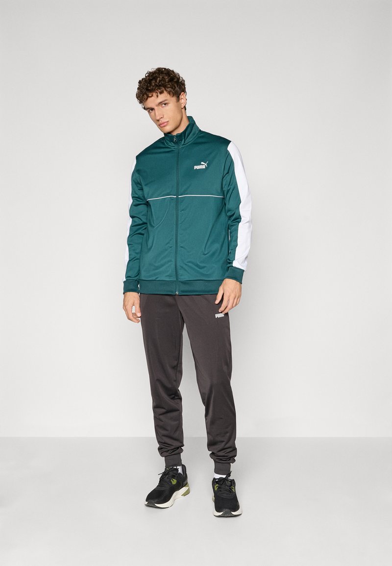 Treningsdrakt med en teal-jakke med hvite detaljer og en logo, sammen med mørke bukser. Laget av glatt syntetisk materiale.