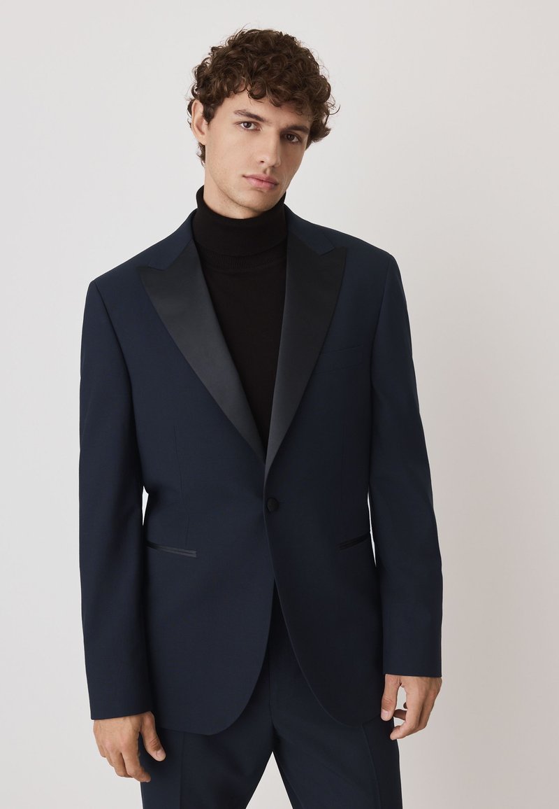 Veste de costume bleu marine avec revers en pointe en satin, fermeture à un bouton et deux poches avant. Portée sur un col roulé noir.