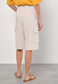 Beige cargo shorts memiliki potongan longgar dengan pinggang elastis, kantong samping, dan tekstur yang halus. Dipadukan dengan sandal cokelat.