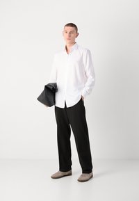 Chemise blanche à manches longues et à boutons, pantalon noir ajusté, tenant un sac en cuir noir. Les chaussures sont beige clair avec des semelles plus foncées. Design minimaliste.