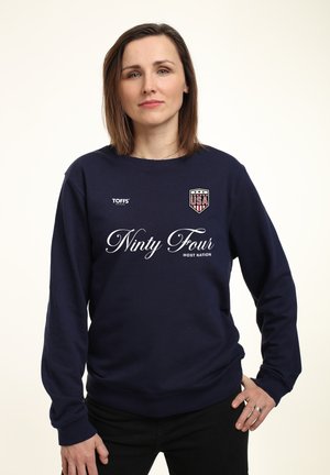 Kvinde med skulderlangt brunt hår iført en navyblå sweatshirt med teksten "Ninty Four Host Nation" og et USA-emblem på brystet.
