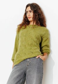 Pull en tricot vert olive avec un motif ondulé texturé. Doté d'un col rond et d'une coupe décontractée. Assorti à un jean taille haute gris.