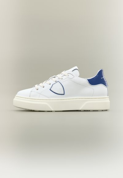 Baskets blanches avec semelle épaisse crème, languette de talon bleue, logo en forme de bouclier bordé de bleu sur le côté, et lacets blancs, vues de profil extérieur.