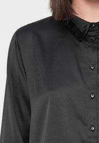 Camisa de seda preta com colarinho clássico, apresentando um detalhe de colarinho em lantejoulas e botões pretos à frente. Textura suave.