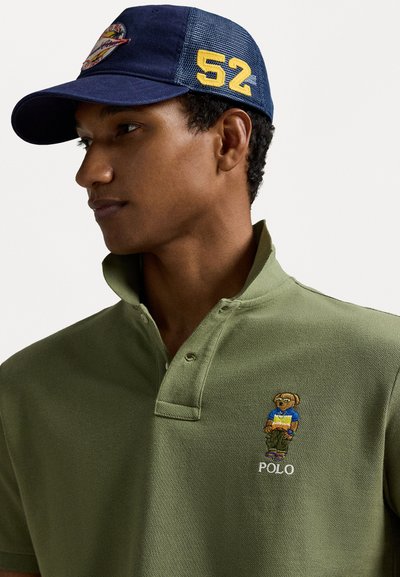 Oliwkowy polo z kołnierzykiem i dwoma guzikami, z wyszywanym logo niedźwiedzia i napisem "POLO". Noszone z granatową czapką, ozdobioną żółtym napisem "52".