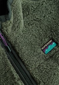 Fleece jack in donkergroen, met een gestructureerd oppervlak, ritssluiting en een logopatch met blauwe en paarse accenten.