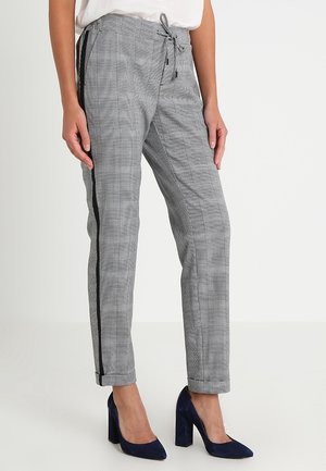 Frau trägt grau karierte, konisch geschnittene Hose mit schwarzen Seitenstreifen und marineblaue Blockabsätze, steht vor einem schlichten weißen Hintergrund.