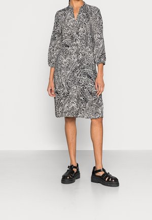 Mujer con un vestido de estampado paisley en blanco y negro hasta la rodilla, con mangas tres cuartos y sandalias negras con tiras gruesas.