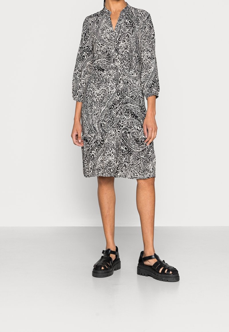 Femme portant une robe noire et blanche imprimée paisley longueur genoux, manches trois-quarts, et sandales noires épaisses à brides.
