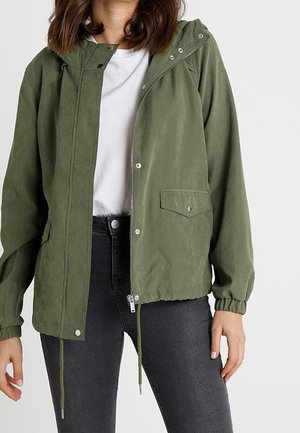 Femme portant une veste à capuche vert olive avec fermeture éclair et boutons-pression, associée à une chemise blanche et un jean gris foncé, les mains détendues le long du corps.