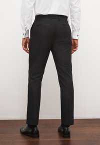 Muž stojí čelom ku stene, oblečený v čierne spoločenské nohavice, bielu košeľu s manžetovými gombíkmi a čierne spoločenské topánky na drevenej podlahe.