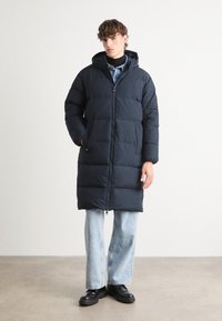 Navy blue puffer coat with a hood, featuring a zip closure and horizontal stitching. Worn over light blue jeans and black shoes.