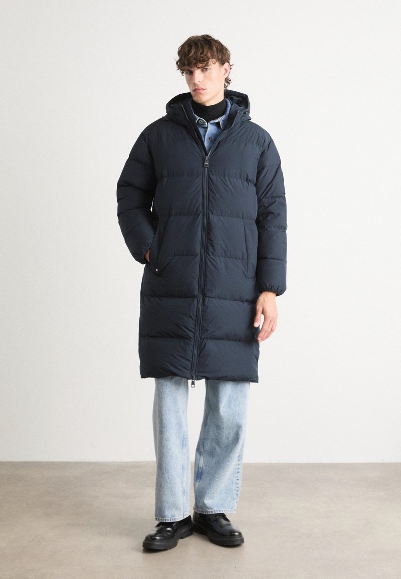 Navy blue puffer coat with a hood, featuring a zip closure and horizontal stitching. Worn over light blue jeans and black shoes.