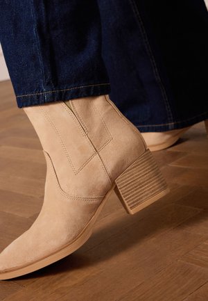 Light beige suede ankle boot with a square toe, sturdy ribbed block heel, and visible stitching details, worn with dark denim pants.