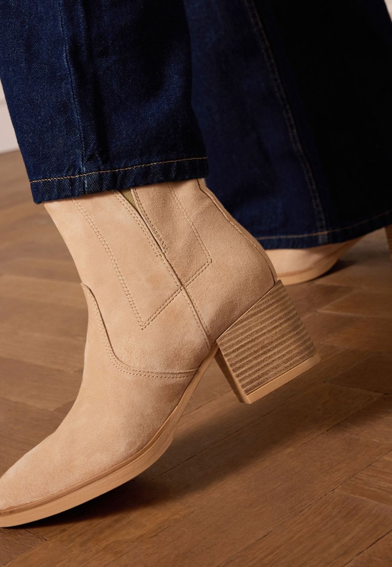 Stivale alla caviglia in suede beige chiaro con punta quadrata, tacco robusto a blocco rigato e dettagli di cucitura visibili, indossato con pantaloni in denim scuro.