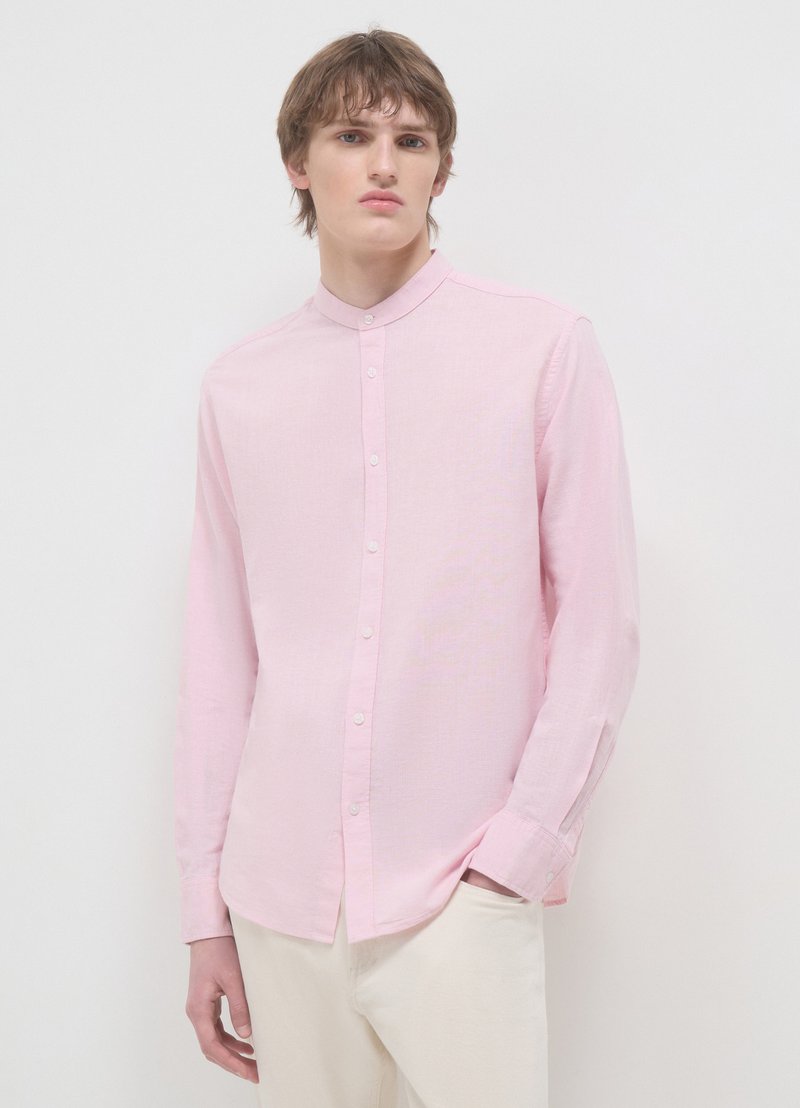 Chemise rose clair à manches longues avec un col mandarin, des boutons sur le devant et un tissu texturé. Portée avec un pantalon de couleur crème.