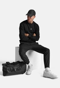 Sort atletisk sweatshirt med logo, matchende joggers, hvide sneakers og en sort duffelbag, alt sammen på en lys baggrund.