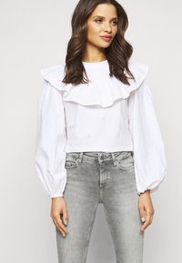 Blusa branca de mangas longas com colarinho frisado e mangas balão, apresentando detalhes de ilhós. Combinada com jeans cinzentos ajustados.