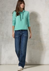 Helles Teal-Hoody mit schwarzen Kordeln, kombiniert mit lockeren blauen Jeans und weißen Sneakers. Glatte Textur, lässige Passform.