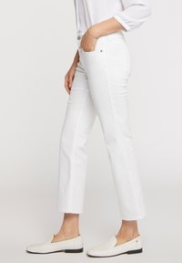 Jeans de mezcla de algodón blancos con corte recto, bolsillos frontales y cierre frontal con botón. Llevados con zapatos tipo slip-on de color beige claro.